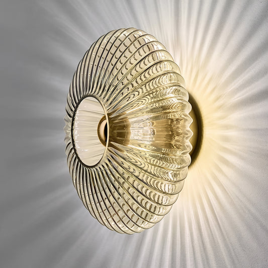 Applique murale en verre nervuré cognac allumée sur platine laiton, rayons de lumière sur un mur gris